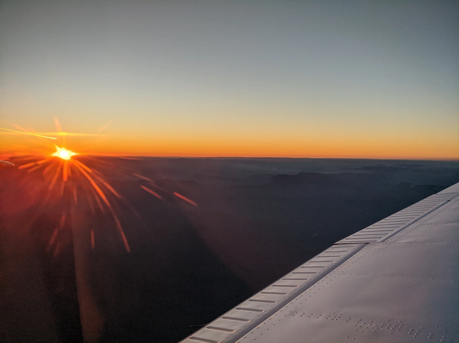 Low sun over distant ridgelines, photographed from an aircraft with its wing in the foreground